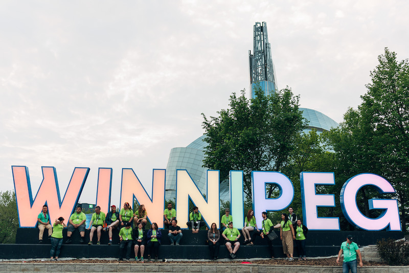 The Winnipeg sign at The Forks lit in rainbow colours with the Canadian Human Rights Museum building behind it.